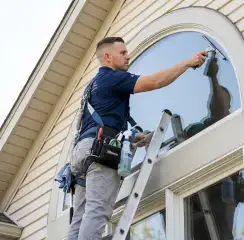 Professional window cleaner on a ladder using a squeegee to clean an exterior arched window, Worcester, MA.